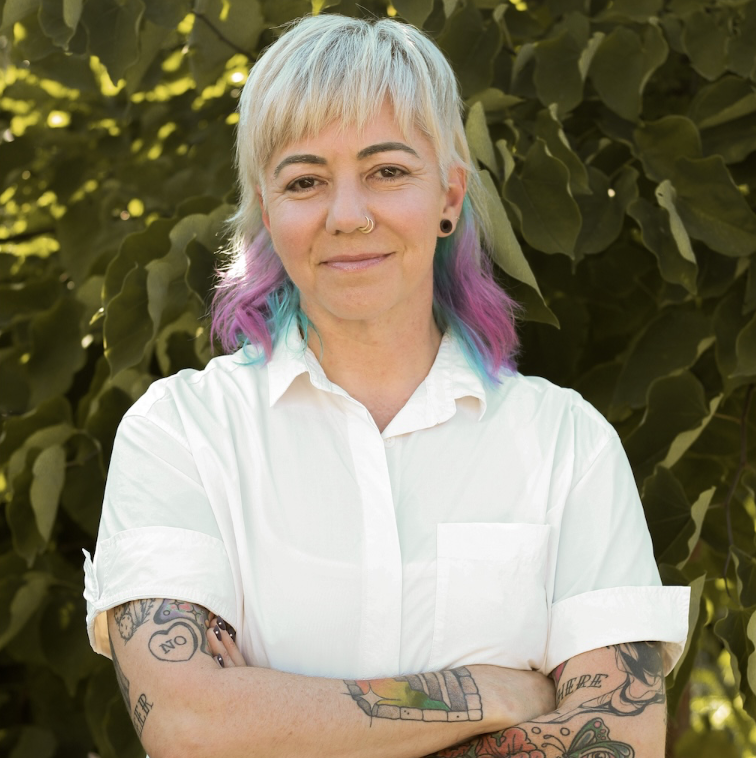 Portrait of Sarah Mark standing outdoors in front of leafy green foliage. They have short platinum-blonde hair with turquoise and purple accents, and are wearing a white short-sleeve button-down shirt. Their arms are crossed, revealing several colorful tattoos on both forearms. They are smiling slightly and looking toward the camera.
