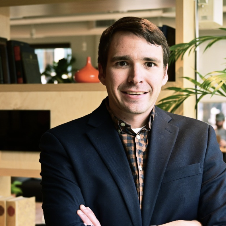 Professional headshot of Mike Kemp standing indoors with their arms crossed. They are wearing a navy blazer over a brown-and-blue plaid button-down shirt. Behind them is a modern office setting with shelving, books, plants, and decorative objects. They are smiling and facing the camera.